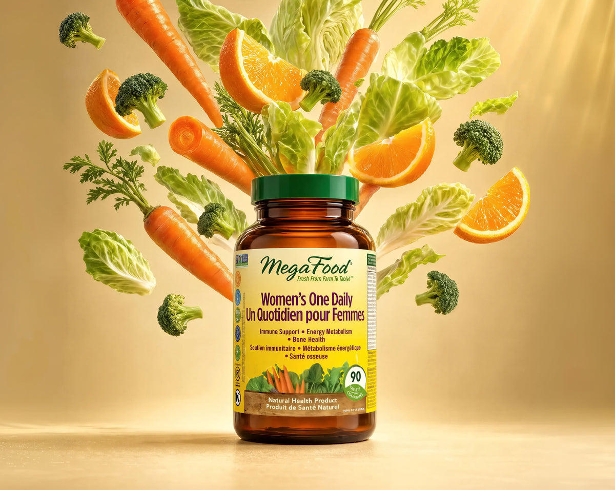A bottle of MegaFood Women's One Daily dietary supplement centered on a warm beige background, surrounded by floating fresh vegetables and citrus slices including carrots, broccoli, cabbage, and orange slices.